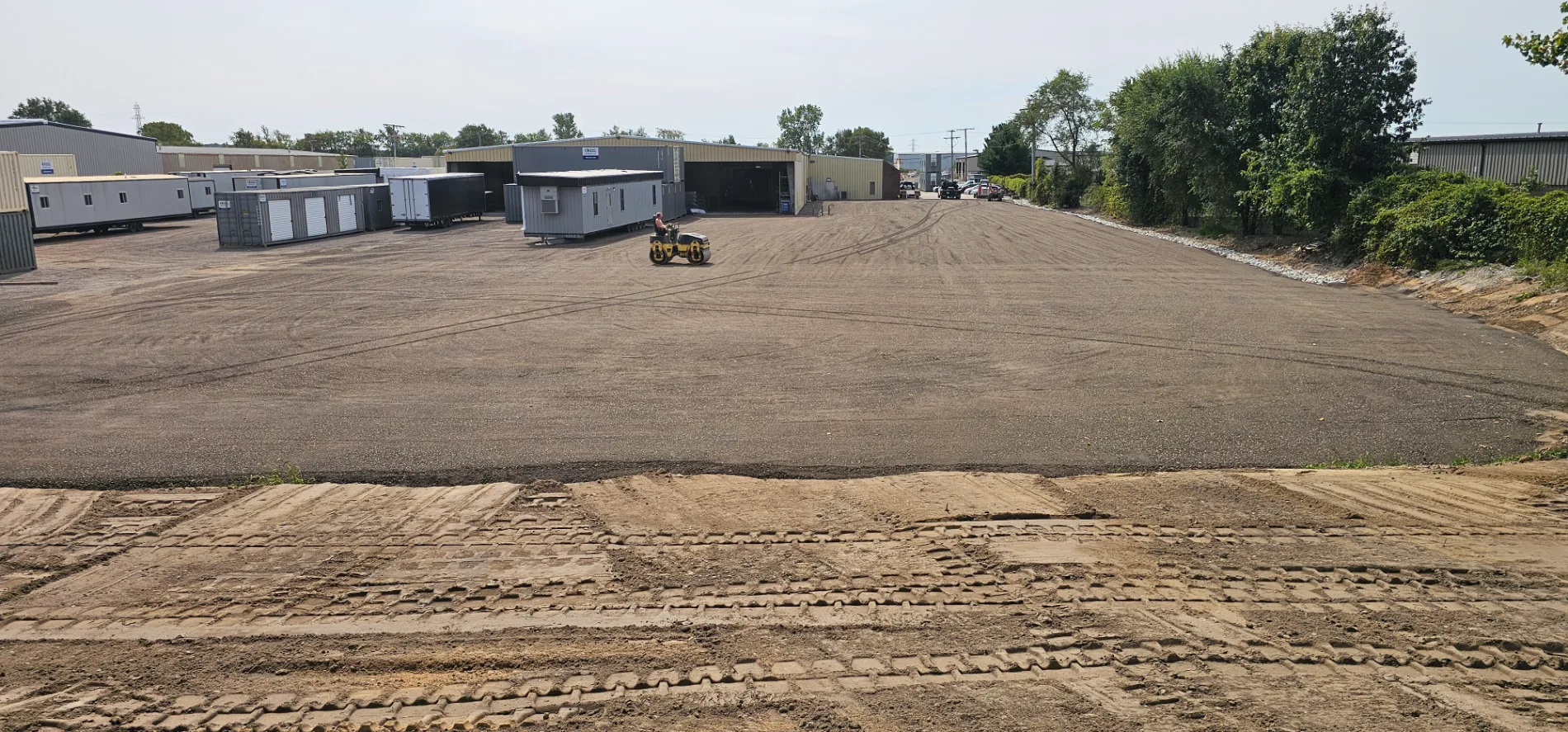 A yellow roller compactor leveling a large gravel lot during a site grading project.